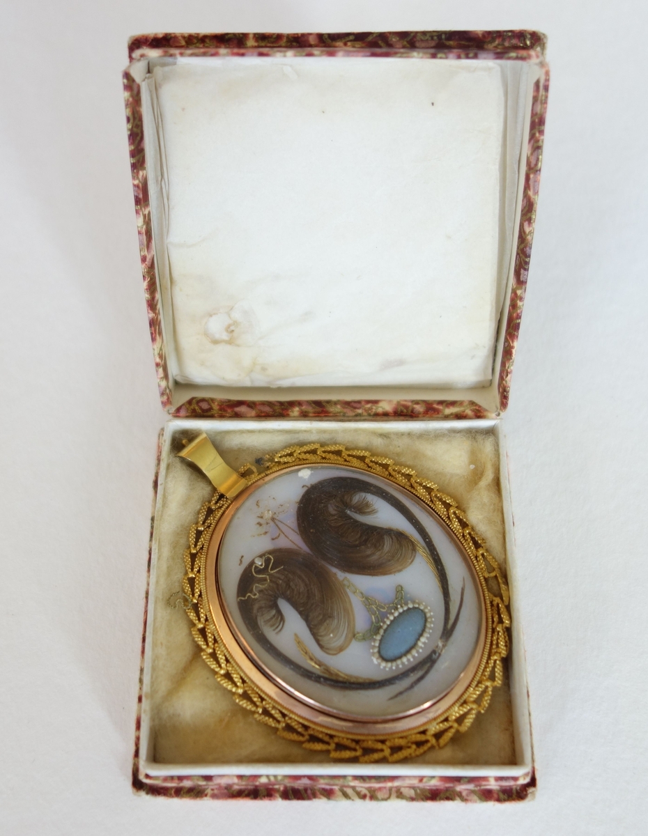 Oval medaljong i gullramme. To mørkblonde hårlokker symetrisk dandert, i midten en blå oval omkranset av små hvite perler. Marmorert etui av papp, fôret med silke i lokket og annet stoff i bunnen.