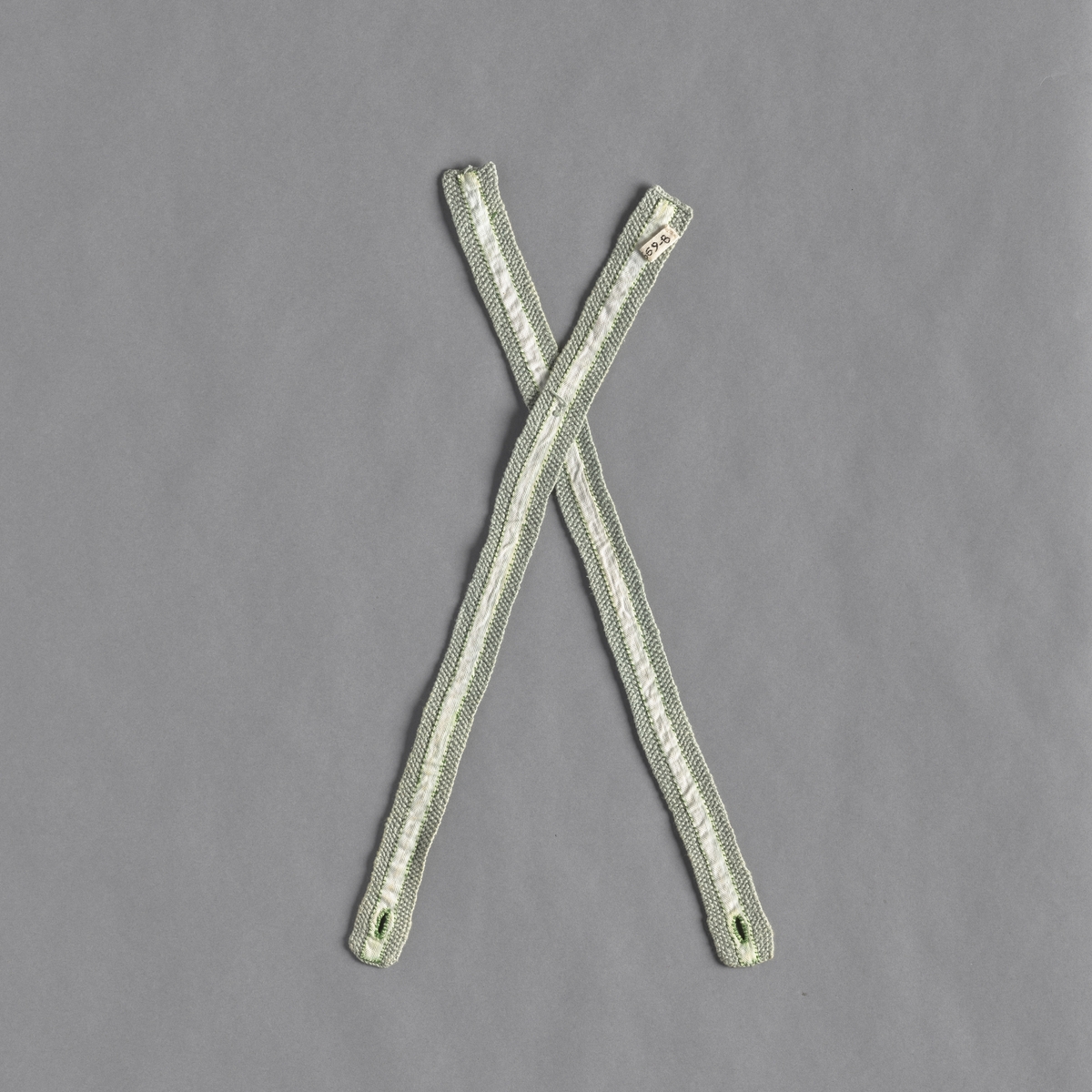 Sele strikket i perlestrikk. To lange strikkede stykker er sydd sammen. Det er sydd et hvit bomullsbånd på stykkene. Selen er i en lys grønn farge.