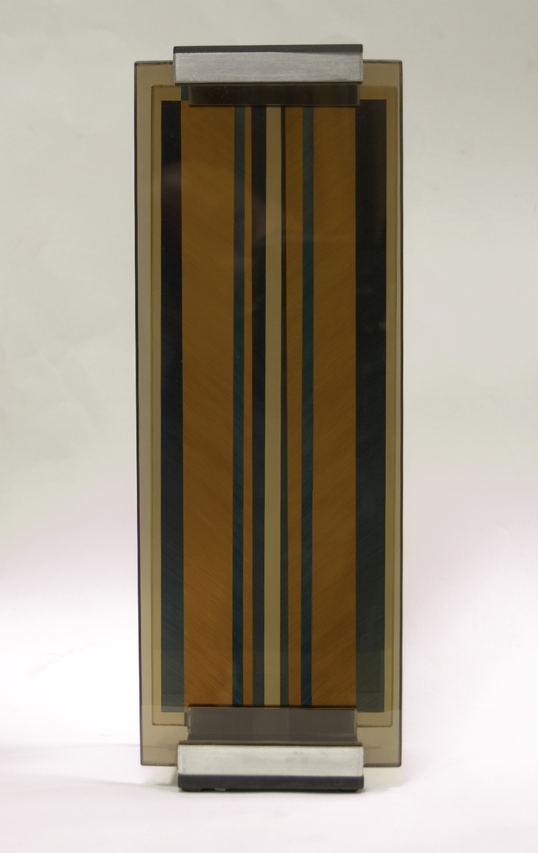 Skulptur i glas, "Optochromi H-4", av Erik H Olson. Mot en svart bottenplatta två horisontella, parallella glasskivor. Genom inlagda genomskinliga segment uppstår vid rörelse spektrala färgbrytningar.
Signerad i botten.