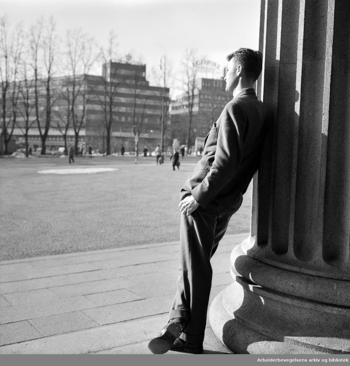 Vår i Oslo. Ung mann lener seg til en søyle på Universitetet. Odd Fellow-gården i bakgrunnen. Foto: 10. Mars 1960.