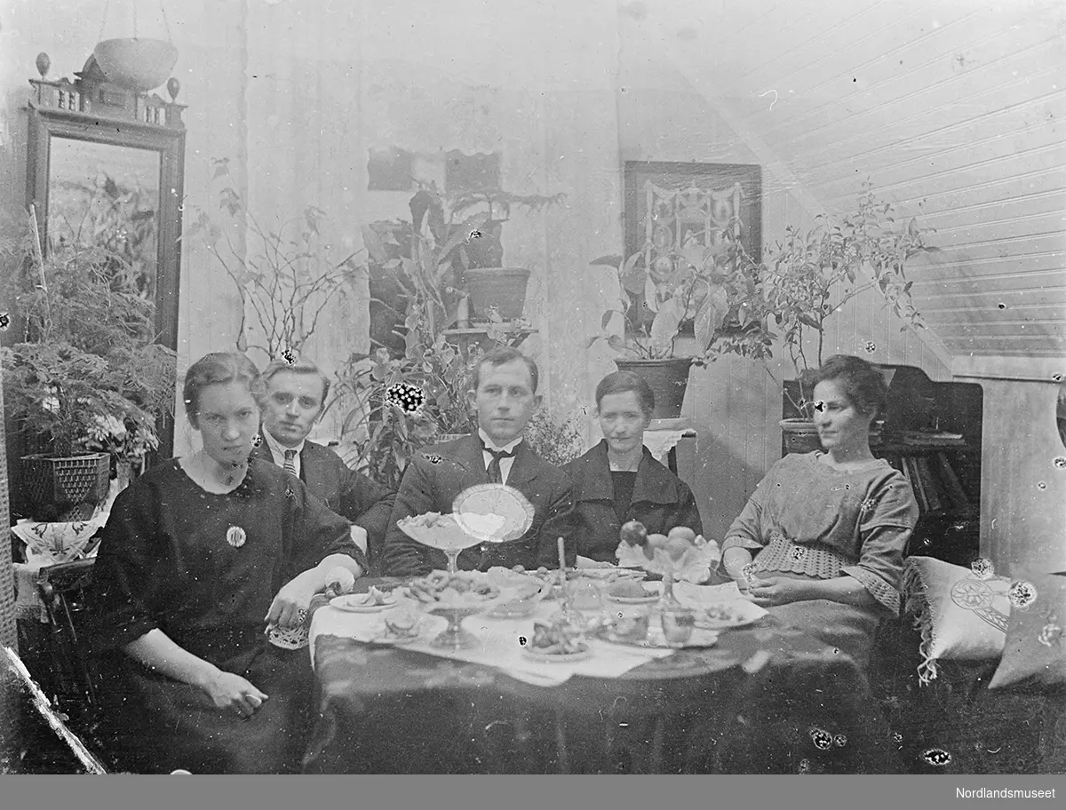 Vi ser tre damer og to herrer sittende rundt et bord i en stue. Paret til venstre er gift. I venstre del av bildet ses et stort speil. Rundt i rommet står blomster og kaktus. På bordet står forskjellige kakefat og en kurv med epler.