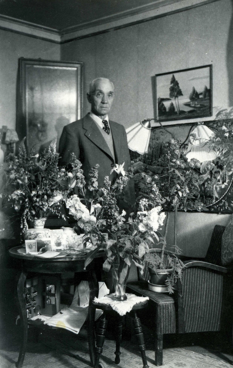 Uppvaktning av Olof Olsson (1883 - 1958) som står omgiven av buketter i bostaden "Alphyddan" Torrekulla 1:25 på Gamla Riksvägen 13, okänt årtal. Han var S-politiker i Kållered samt skötte ABF-bilblioteket.