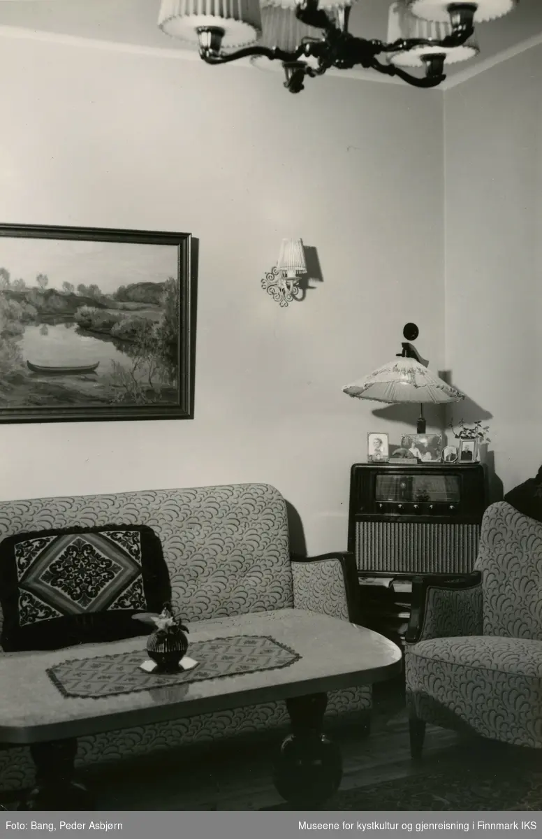 Bildet viser en del av en stue på 1950-tallet. Det er mest sannsynlig stua til direktørboligen i bygget til Finnmark Fylkesrederi og Ruteselskapet på dampskipskaia i Hammerfest.
Man ser et stuebord som er pyntet med duk og blomstervase. Til venstre er det en sofa med pute. Over sofaen hender det et landskapsmaleri med en elvebåt og en vegglampett til høre for den. Til høyre står det en passende lenestol.  I hjørnet mellom sittemøblene er det plassert et radiobord med radio og en gulvlampe bak det. Opp på radioen står det fire innrammede fotografier og en liten isbjørnskulptur. Øverst i bilde ser man en femarmet elektrisk taklampe, på gulvet ligger det et teppe.