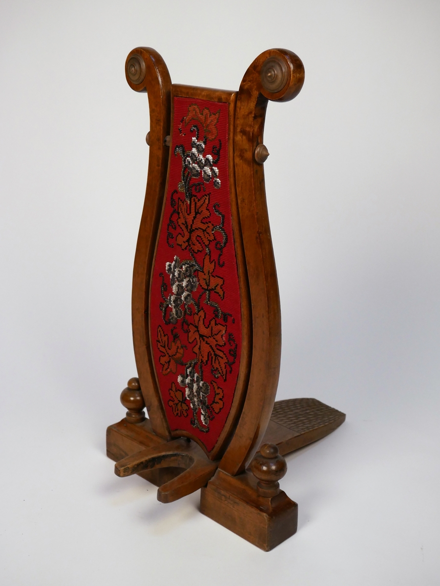 Stövelknekt bestående av en lyrformad träställning med en mittplatta av röd textil prydd av broderi.