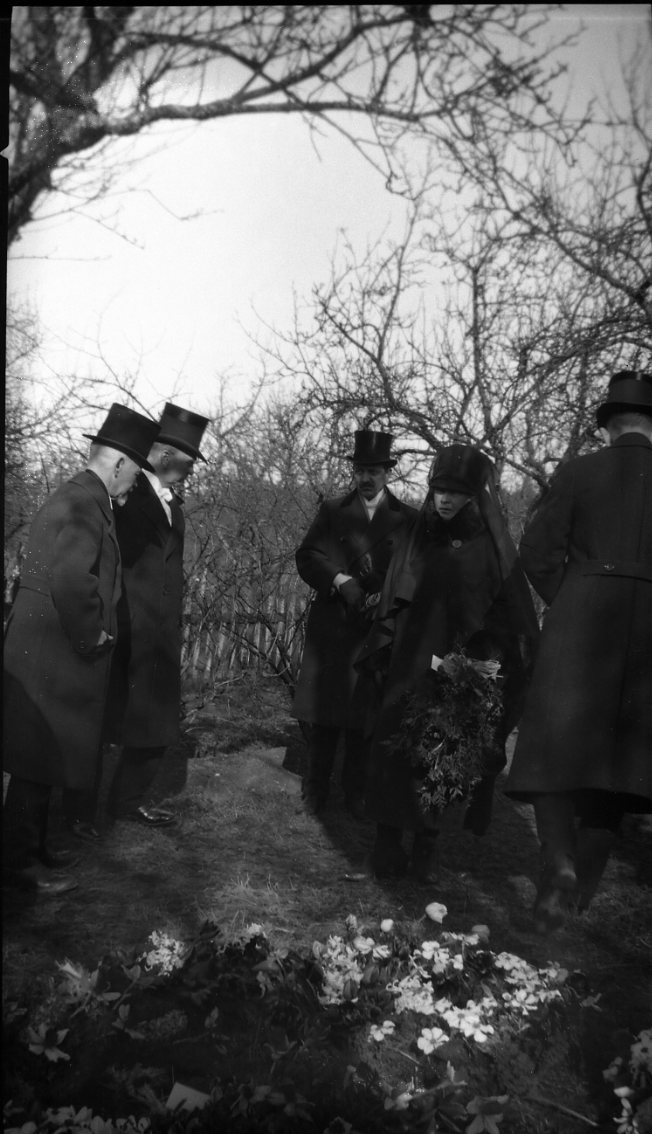 En kvinna med sorgflor och fyra män med hög hatt står samlade vid en grav.