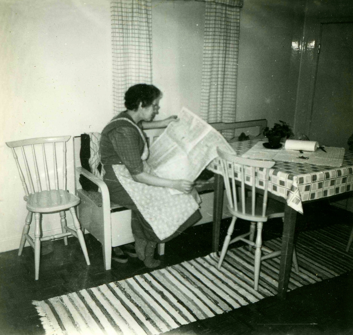 Margit Bengtsson (1901 - 1998) sitter och läser i sitt kök på första våningen i Heljered Sörgård "Skräddarns" 1950-tal. Margit och maken Erik brukade och ägde gården som senare revs. Sedan år 2013 ligger Heljereds förskola på platsen.