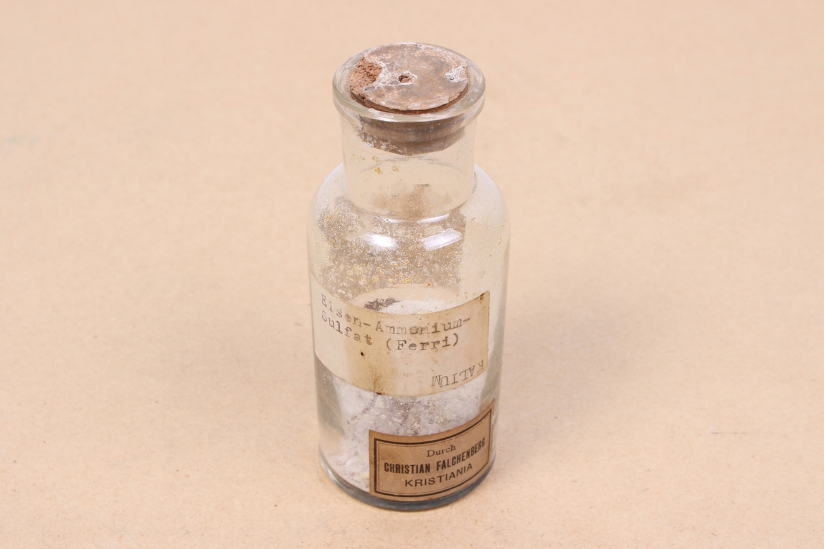 Kjemikalieflaske av klart glass, med stopper av kork. Inneholder ammonium jern(III)-sulfat, en type alunsalt som har blitt brukt blant annet i garving.