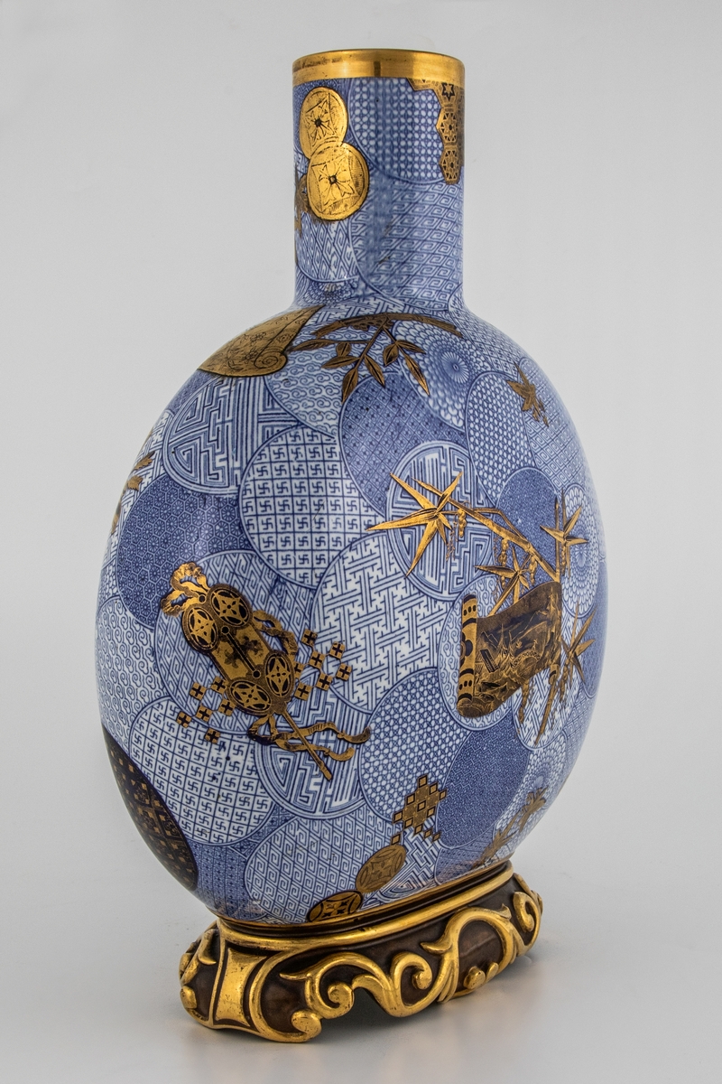 Stor vase med sylinderformet hals og flattrykt kuleformet korpus på kraftig fot med forgylt rankedekor. Mønstret sirkeldekor i blå underglasur, samt bambus, kostbare gjenstander og sirkelmønstre i mørkere blått og gull.