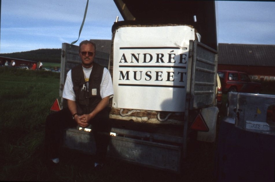 Man sitter i en släpvagn med öppen kåpa. Bredvid honom står en ballongkorg med texten Andréemuseet på.