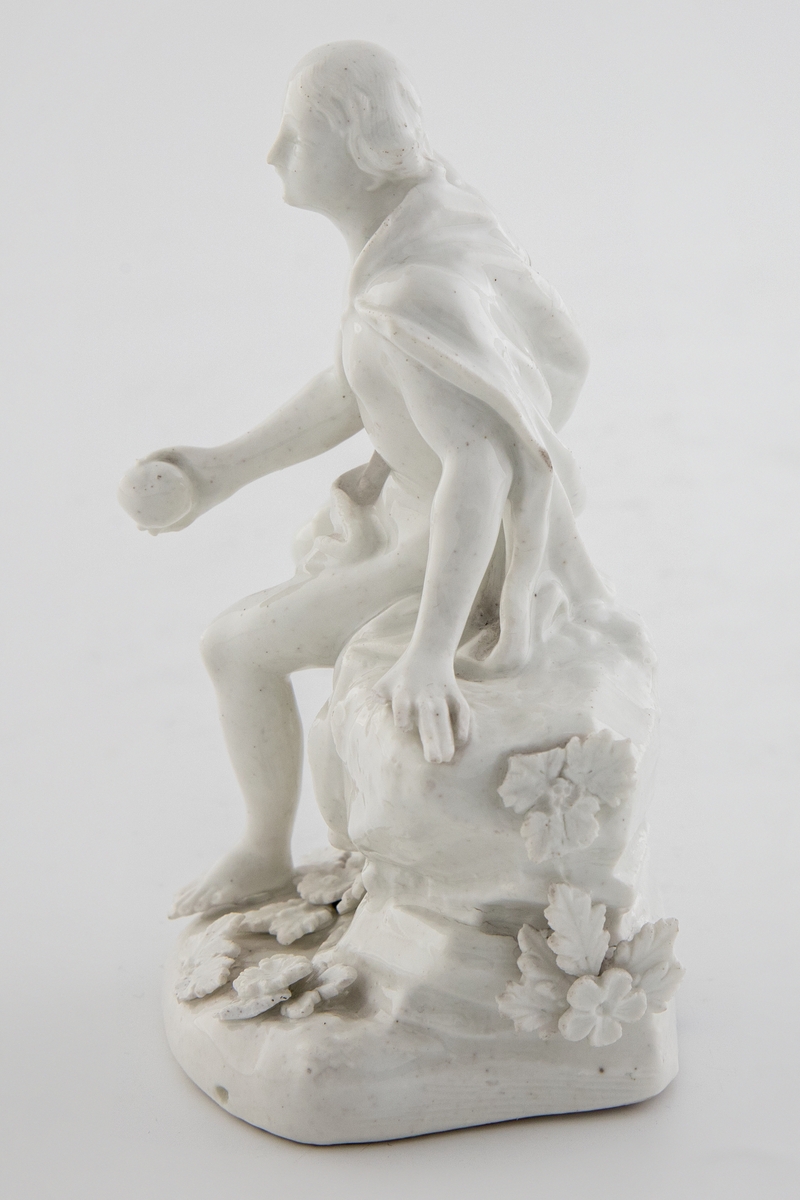 Hvitglasert, umalt porselensfigur med en ung mann sittende på en klippe. Figuren holder et eple i høyre hånd og identifiseres som den trojanske prinsen Paris fra gresk mytologi. Sokkelen er dekorert med blomster og blader.