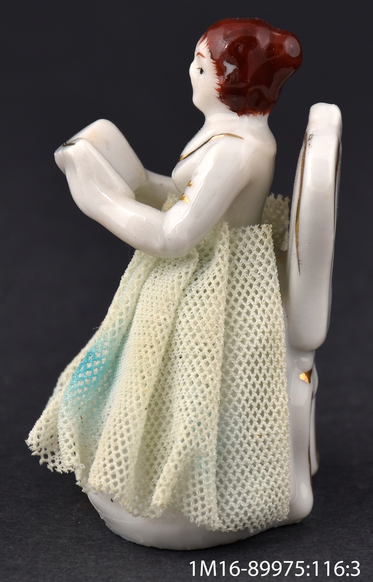Leksak av porslin, i form av en sångerska som sitter på en stol i vitt och guld. Sångerskan håller en bok i sina händer. Kjolen är gjord av veckat tyg.
