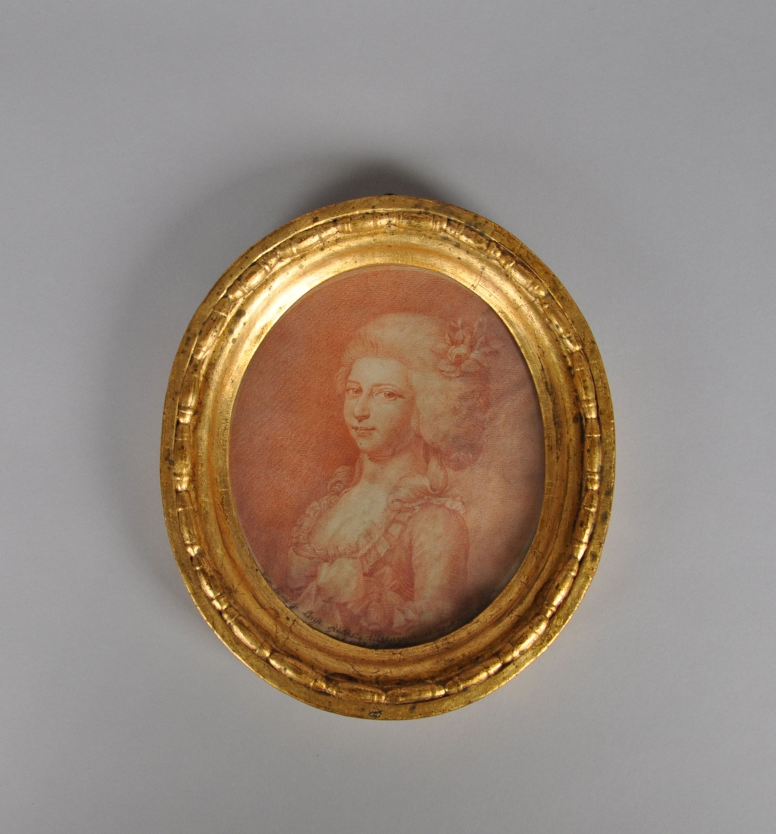 Trykk av jente i barokkjole, med blomsterpryd i håret. Trykket har rødfarge. Stående oval form, med gullbelagt ramme. Portrett av Prinsesse Louise Augusta av Danmark.