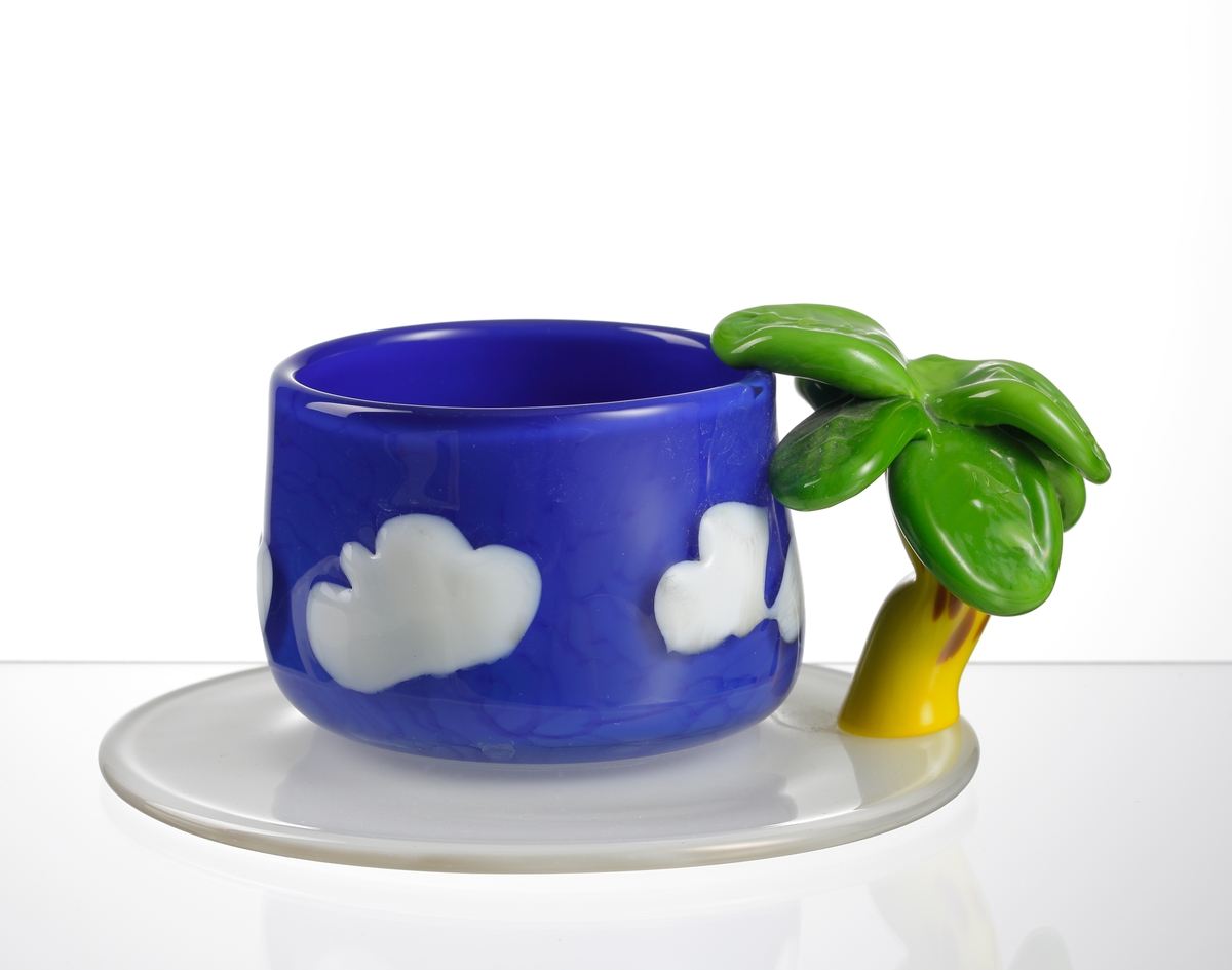 Kopp/tekopp på fat, "Palm Tree Cup", formgiven och tillverkad av Paula Bartron.
Koppen är blå med vita "moln" och grepen består av en palm med gul stam och gröna blad. Fatet är vitt.