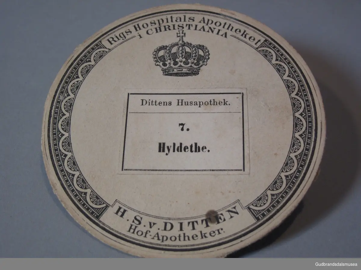 Rund plakett i papp med varebetegnelsen: Dittens Husapotek. 7 Hyldethe.