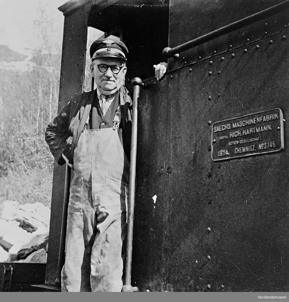 Mann med uniformslue, briller og kjeledress står i døråpningen på et lokomotiv. Lok og lokfører på Sulitjelmabanen, lokomotivfører Hansen.