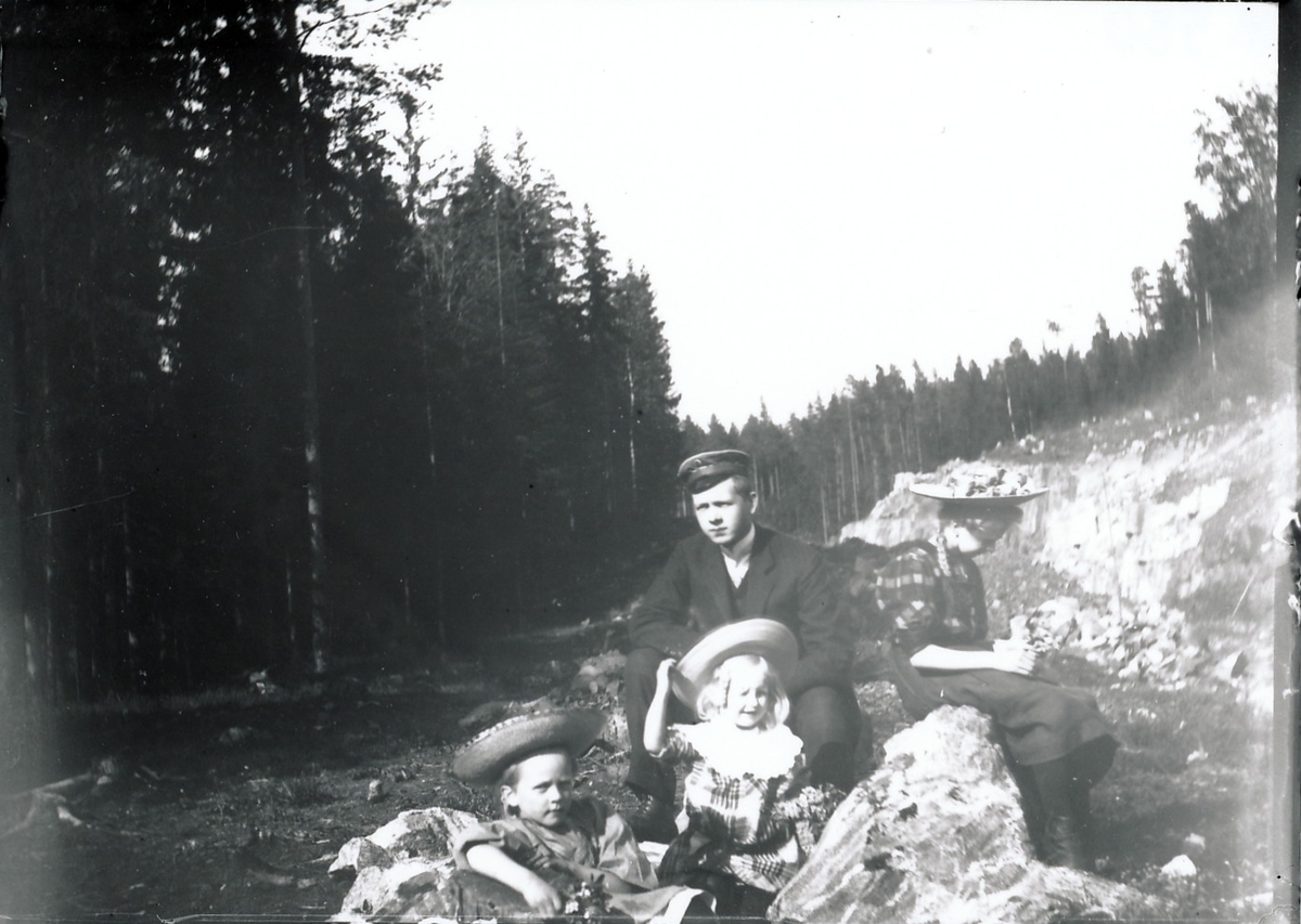 Ung gutt og tre jenter sitter på steiner sannsynligvis i forbindelse med veibygging. Jentene har stråhatter, gutten skyggelue. Til høyre er en bergvegg, som det ser ut til å være sprengt ut fra, og til venstre er det skog.