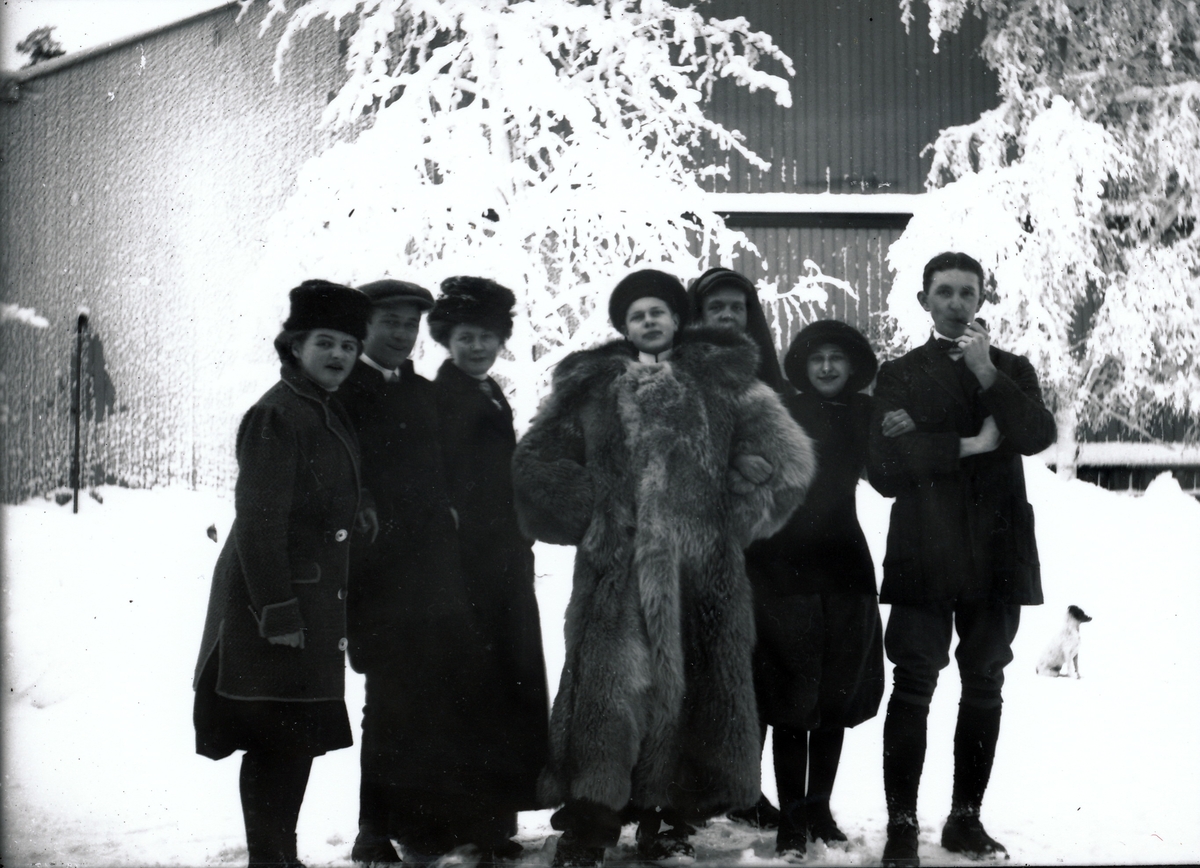 Tre unge kvinner og fire unge menn i gårdsplassen på bestyrerboligen ved Kistefos. I bakgrunnen er den store arbeiderboligen, Boligen. Mannen i midten er iført sid ulveskinnspels. To av kvinnene i knekorte, posete skjørt eller bukser.