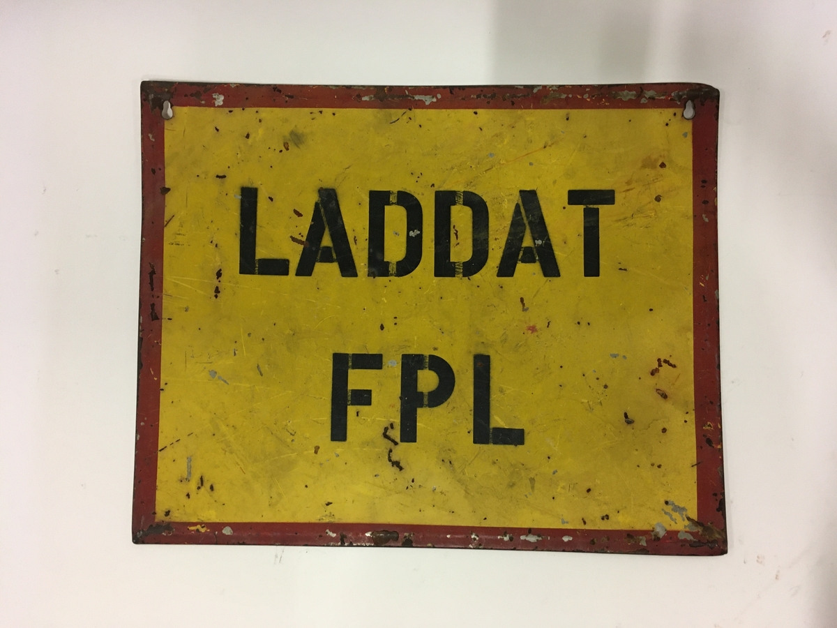 Plåtskylt "LADDAT FPL". Svart text mot gulbotten med röd ram.
