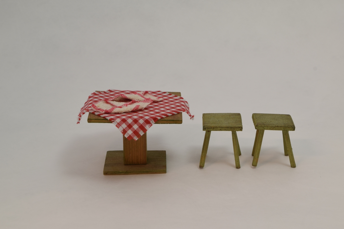 Kjøkkenbord med to krakker i tre. På bordet ligger det en rødrutete duk og to servietter. Møblene er laget av polske krigsfanger og tekstilene er tilført av tidligere eier.