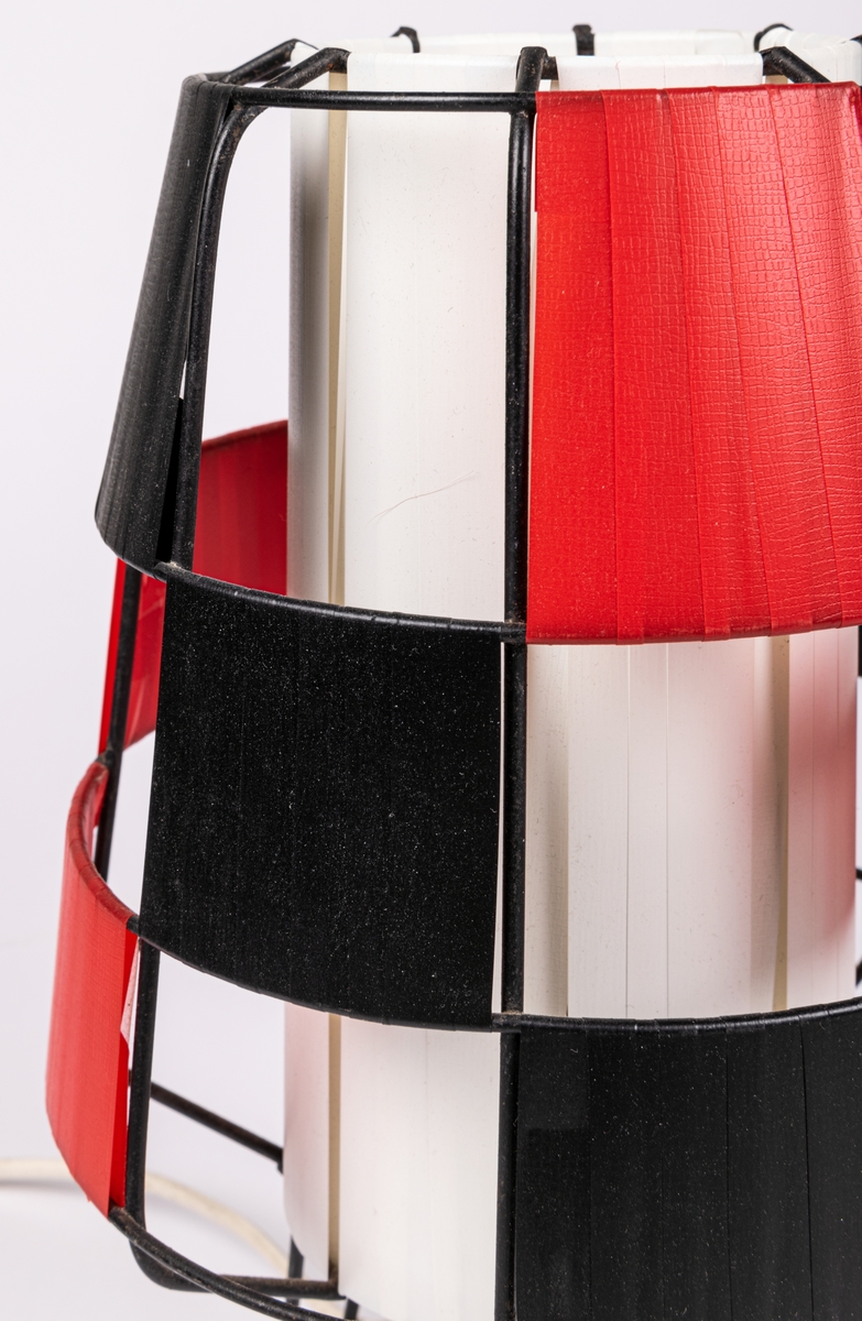 Bordslampa, svart plastklädd metallställning. Plastremsor trädda i rutmönster i svart, rött och vitt. 
Raketformad.