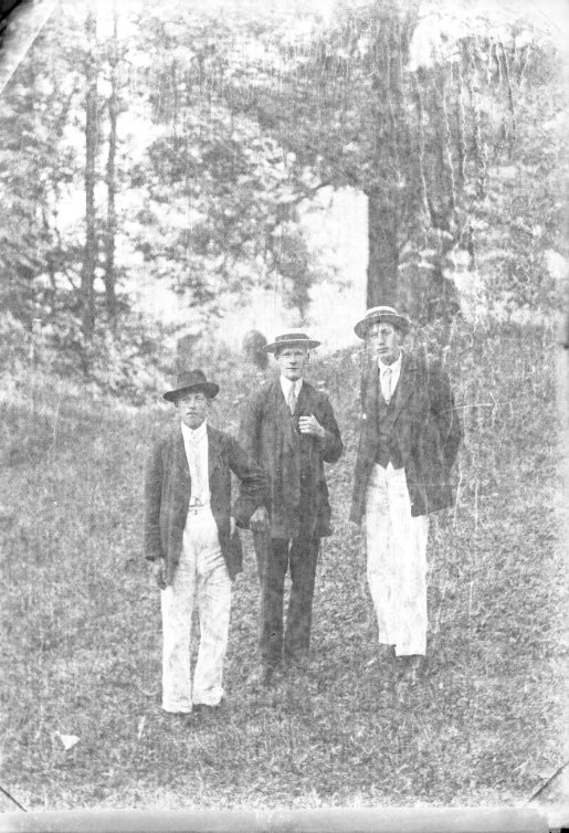 Tre unga män med hatt står på en grässlänt. Två av dem har ljusa byxor.