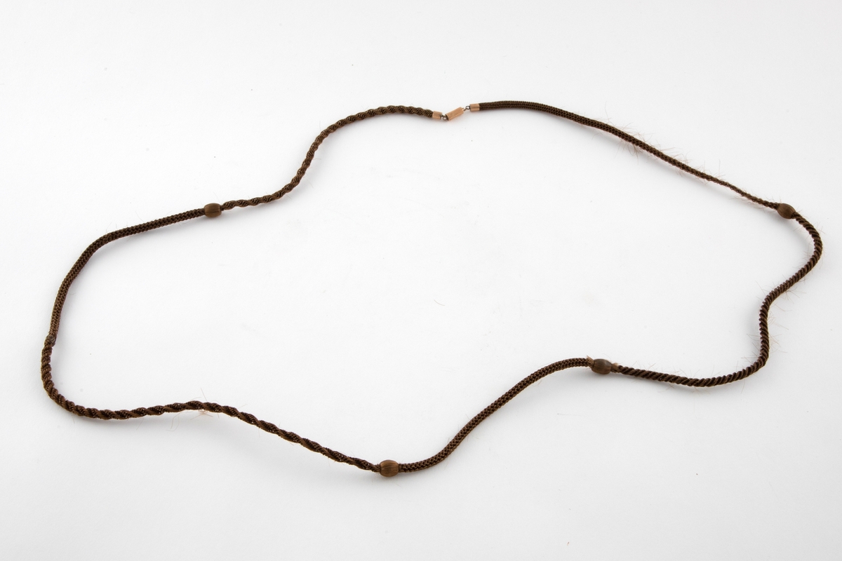 Halssmykke i brunt hårarbeid med lenke av metall som kan åpnes og lukkes i nakken. Smykket er delt inn i seks partier av ulik fletteteknikk. I overgangene mellom partiene er håromspunne treperler, i alt 4 stykker. det ser ut til at en mangler.
