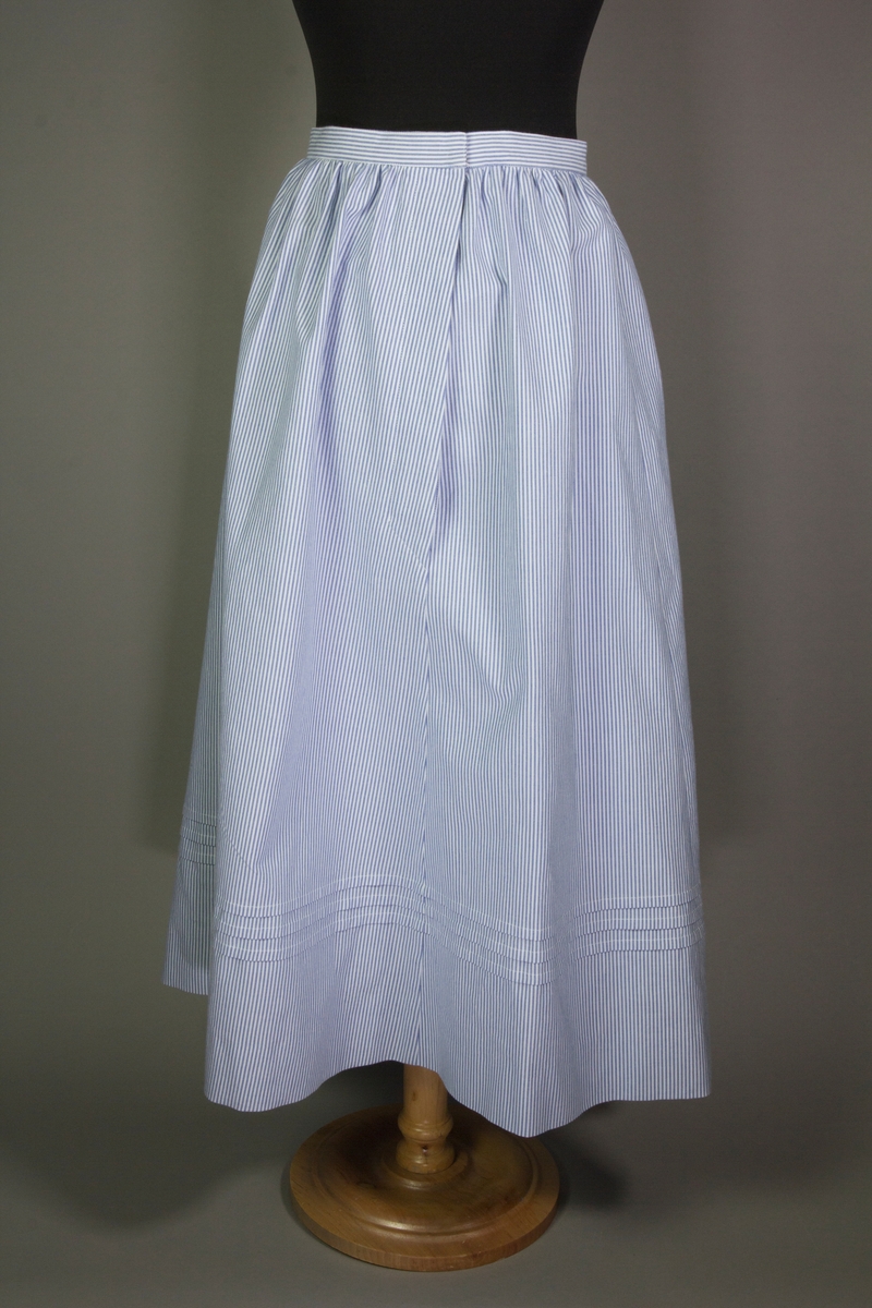 Kjol till tvådelad dräkt av vit och blårandig bomullsväv. Kjolen är hellång med sprund i sidan och tre uppvikta veck.