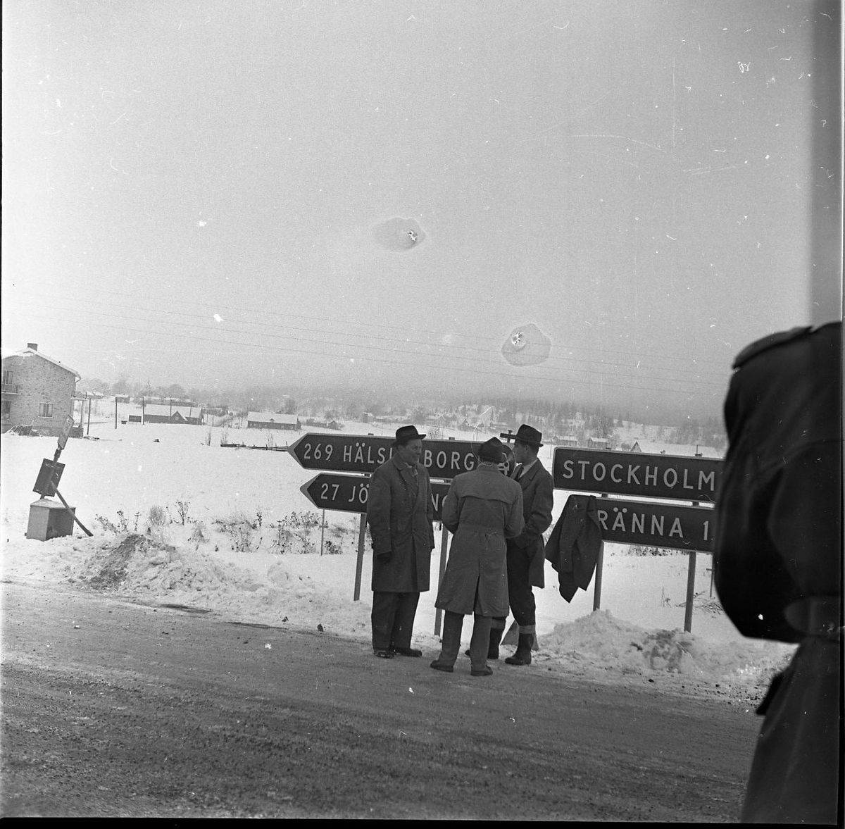 Tre män med hatt och rock står och pratar framför fyra vägskyltar. Vägskyltarna visar avståndet till Jönköping, Helsingborg, Stockholm och Gränna. I bakgrunden syns flera av gårdarna i Ölmstad by. Huset till vänster med fasadtegel är Thuvanders Speceriaffär.