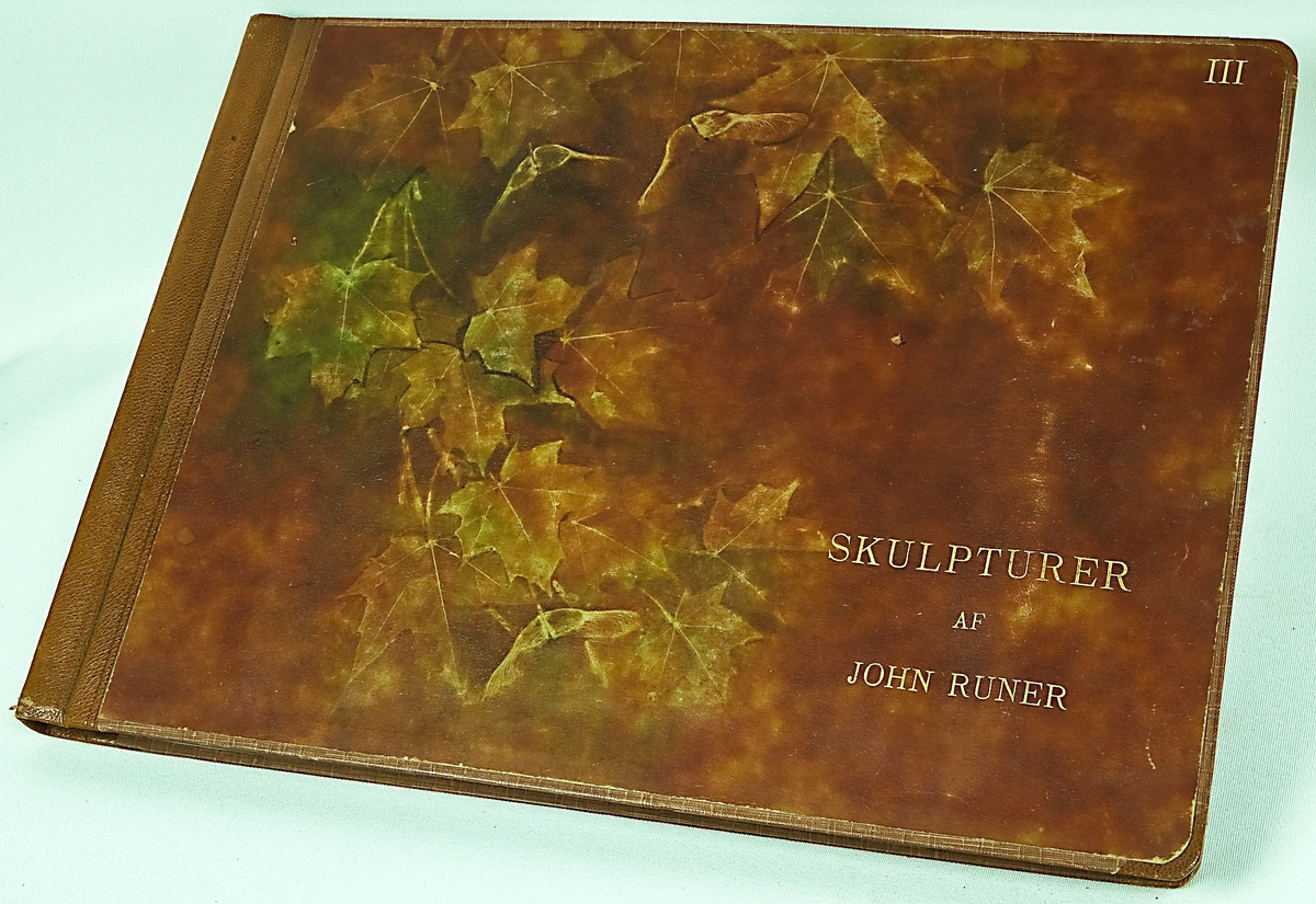Fotoalbum innehållande skulpturer av John Runer, inbunden läderpärm med pappsidor. På framsidan blad och fågelmotiv samt texten "Skulpturer af John Runer III".