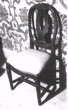 En av tre stolar i gustaviansk stil, ursprungligen klädda med korsstygnsbroderat tyg. Stolarna är dekorerade med snidade blommor i ryggkrön och sarg. Benen är svarvade med utskurna kannelyrer. Ryggbrickan är genombruten och balusterformad. 
Stolen har ursprungligen varit vitmålad med förgyllda ornament.