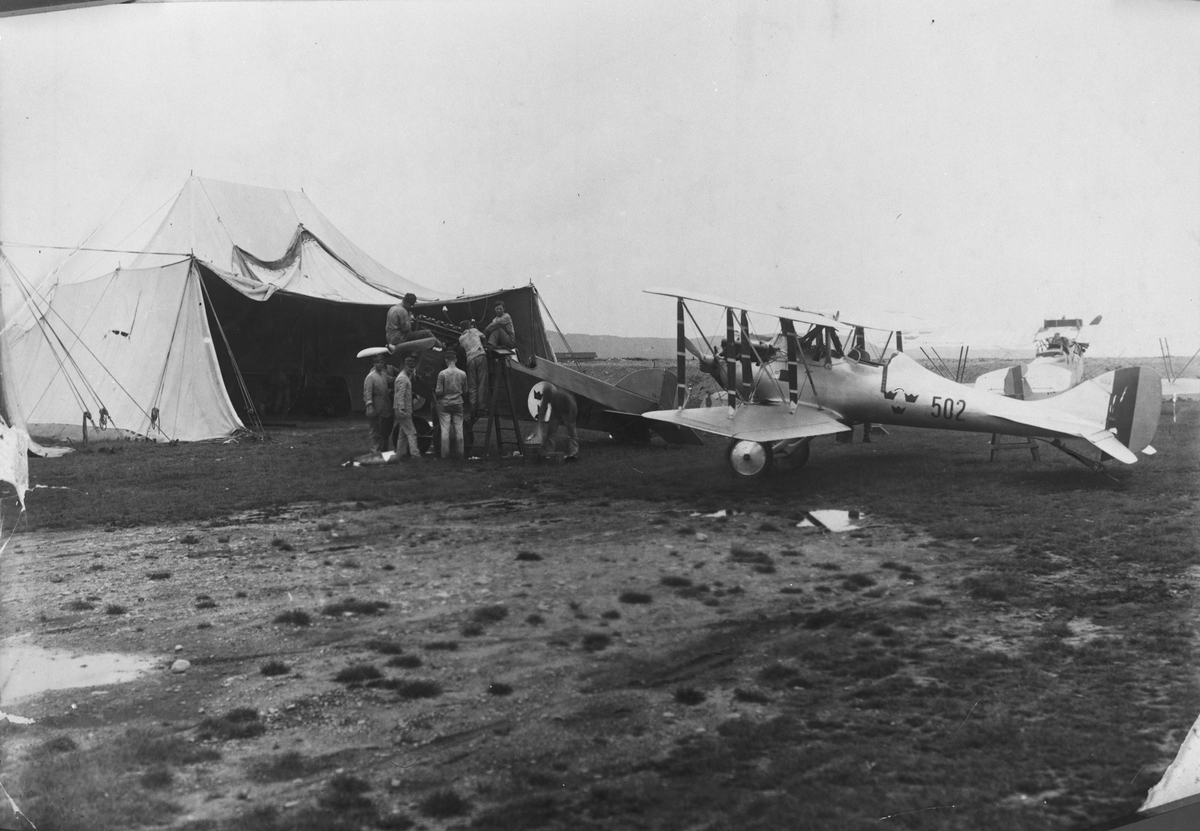 Tekniskt arbete med militära flygplan framför en tälthangar på flygfältet på F 5 Ljungbyhed, 1929-1930. Närmast tältet arbetar åtta flygtekniker med flygplan J 1 Phönixjagaren utan vingar. Därbakom står flygplan J 2, nummer 502. I bakgrunden syns ett flygplan Ö 4 Phönix Dront.