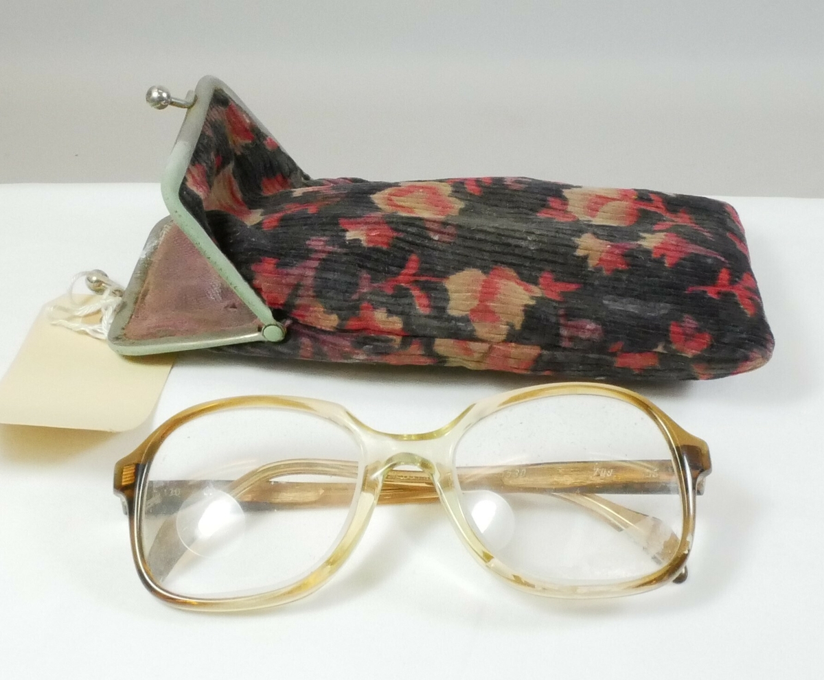 Rektangulær pose i tekstil brukt til brilleetui. Etuiet har kordfløyel med svart bunn og blomstermotiv i gult og rødt, på den ene siden er det en metallskinne med en lukkemekanisme. Inne i er det rosa for. Brilleetuiet inneholder en brille med innfatning i brunt og gult, to brilleglass. 

Påskrift, brille:
METZLER GERMANY