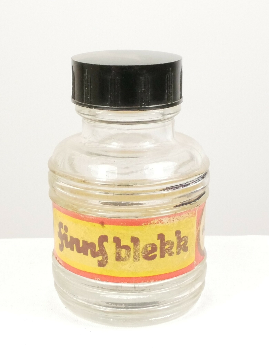 Rund klar glassflaske som smalner inn i en tut som er tettet med en svart plastkork. Rundt flasken er det en papiretikett. 