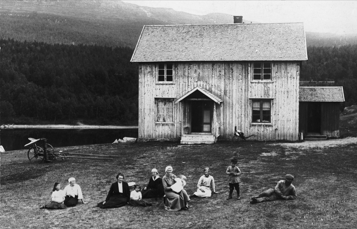 Familie sittende på gårdstunet foran et hus.