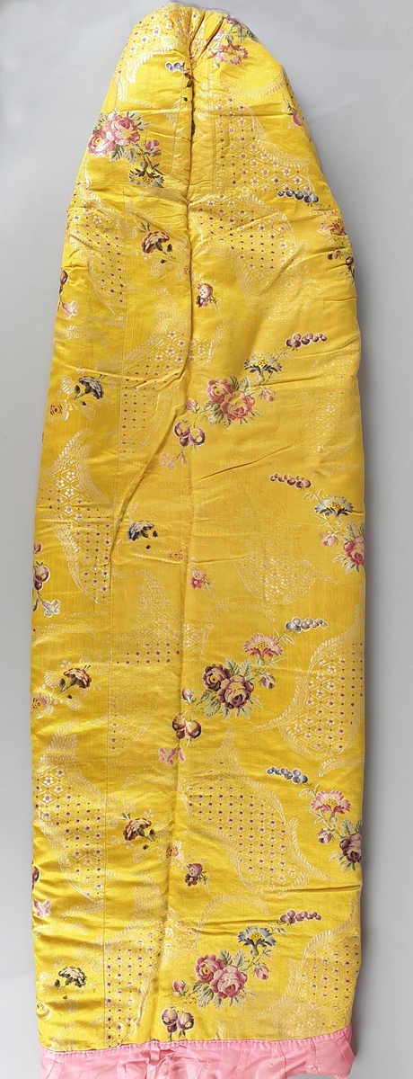Dåpsveip i gul silke med brosjerte blomster, pyntet med rosa rysjekanter. Fór i beige og rød kattun. Lappet i fóret. Dåpssveipet er håndsydd.