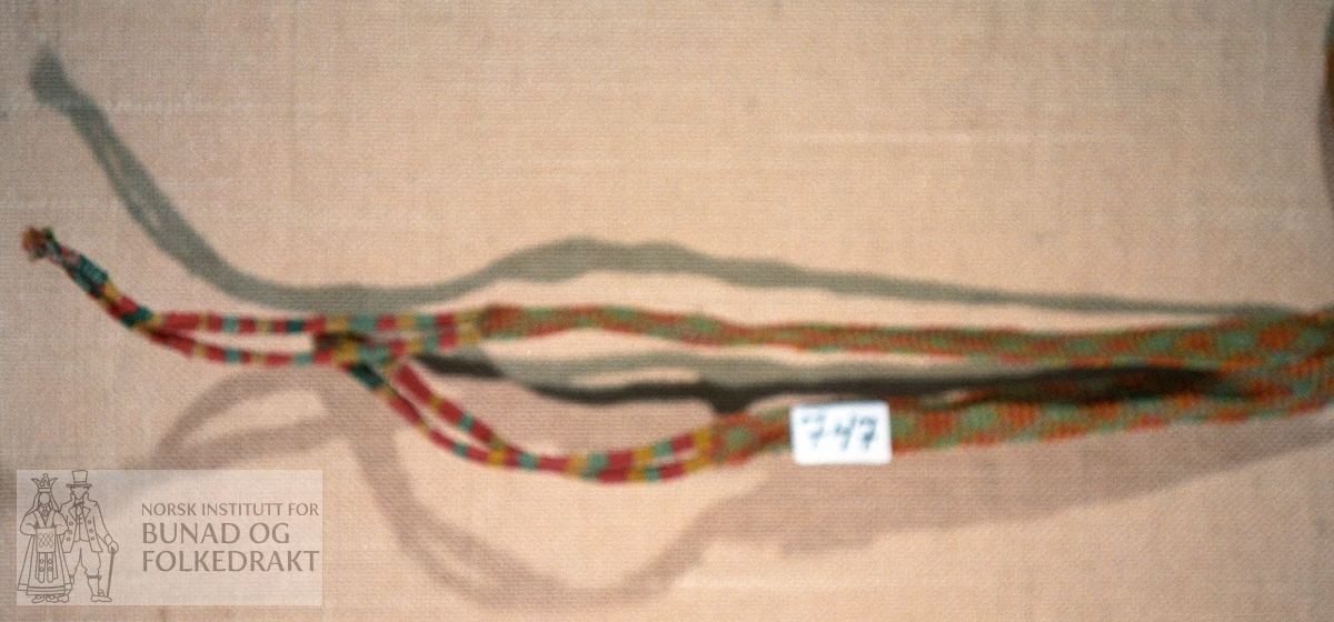 Fingra band av rødt, blågrønt, gult og sauekvitt ullgarn.  I ene enden 2 små tvunnede fletter.  Andre enden 2 fletter med stoppesøm.  L.: 170 cm + 20 cm fletter.  Br.: 2,5 cm.