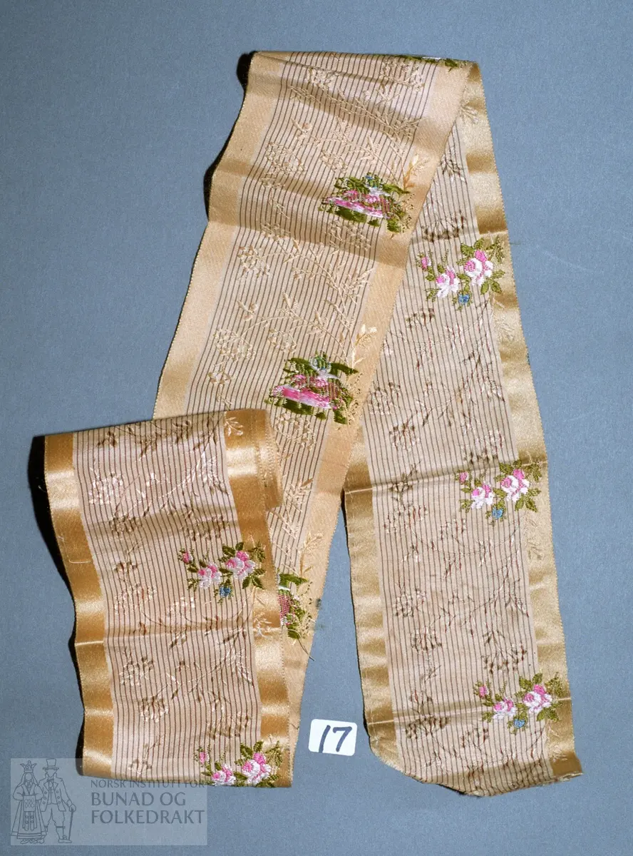 Bredt stripet silkeband i kvitt, gulbrunt og mørk brunt med mønstervevde tynne greiner i rapporter. Brosjerte blomstergreiner ut til ene sida i kvit, rød, grønn og blå silke. Bredde: 7,8 cm. Lengde: 296 cm.
