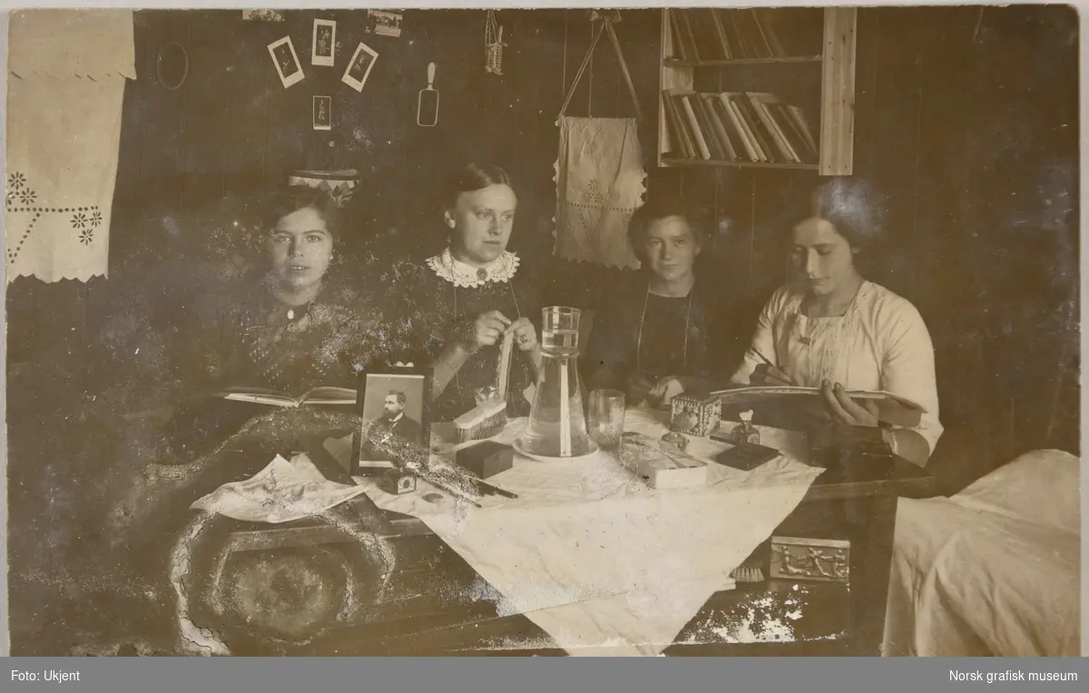 Postkort med bilde av fire kvinner. De sitter rundt et bord og leser, hekler, syr og tegner. På bordet står et bilde av en mann. Det er også børster, penner og blyanter, blekk, en vannkaraffel og et skrin.