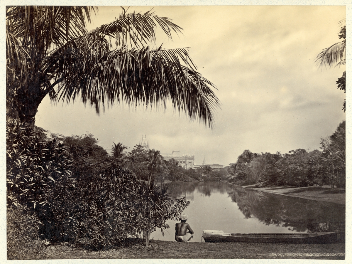 Bilden visar Calcutta äldsta parkanläggning Edens Garden. I förgrunden syns en man bredvid en båt som tittar ut över en vattenyta. I bakgrunden ser man byggnaden av Calcuttas högsta domstol.