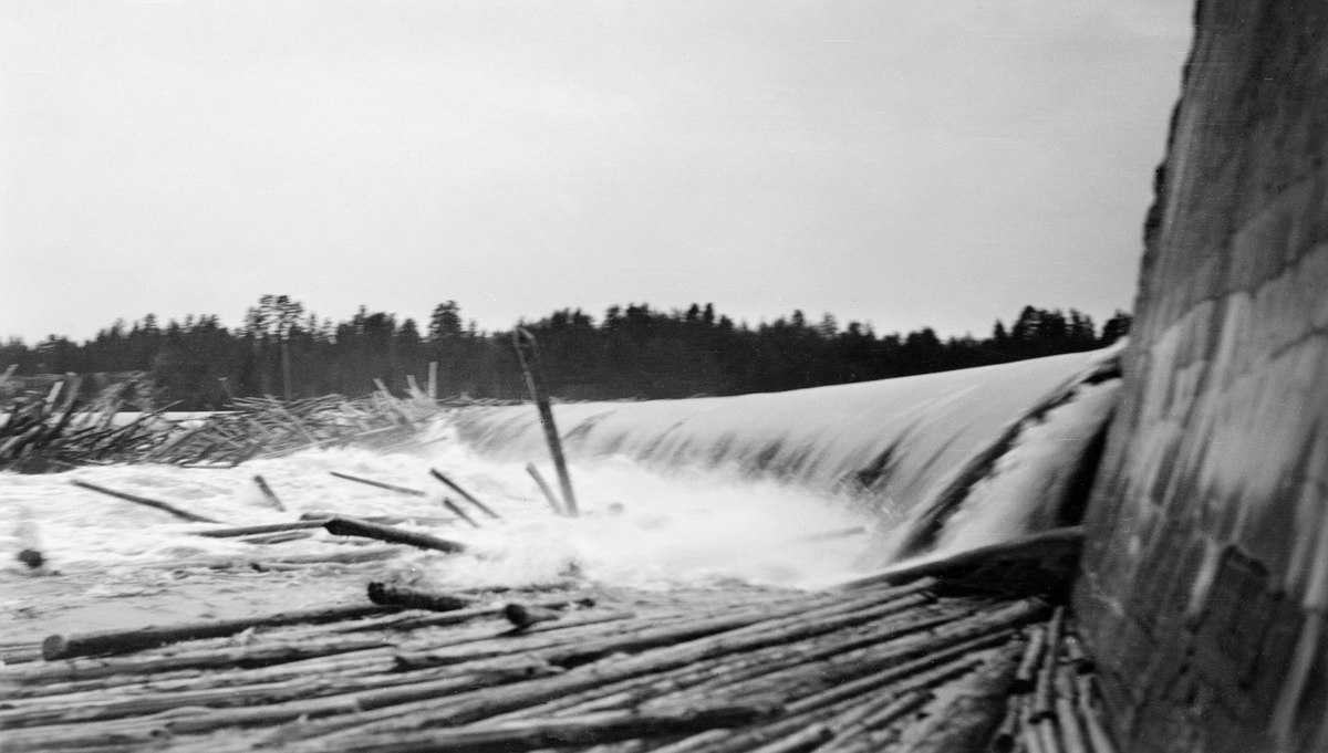 Tømmerfløting ved Skjefstadfossen i Glomma, antakelig sommeren 1933.  Fotografiet er tatt fra en posisjon i le av det høye og kraftige landkaret som forbandt den drøyt 300 meter lange, støpte damkonstruksjonen med vassdragets østre bredd.  Da dette bildet ble tatt rant det vann over både den østre delen av den buete damkrona, som var den laveste, og den midtre, som var en aning høyere.  På begge steder hadde det bygd seg opp hauger av fløtingsvirke, antakelig fordi tømmeret falt fra dammen og ned i et parti der elvebotnen var steinete og ujamn.  Det å løsne slike tømmervaser, mens vannføringa ennå var høy og mulighetene for videre fløting var gode, var farefullt arbeid for involverte mannskaper og en kostnadsdrivende faktor for Kristiania Tømmerdirektion.  Følgelig er det grunn til å anta at fotografiet ble tatt for å dokumentere slike vanskeligheter overfor kraftutbyggerne, som hadde plikt til å holde andre aktører med hevd på bruk av vassdragets skadesløse.

I 1933 var det innmeldt 2 763 841 tømmerstokker til fløting i den delen av Glommavassdraget som ligger nord for Skjefstadfossen. 32 646 av disse stokkene - 1,2 prosent - nådde ikke fram til hovedlensa ved Fetsund i nordenden av Øyeren. Det tømmerhaugene som hadde pakket seg sammen her ved Skjefstadfossen ble sannsynligvis revet, og det var antakelig få av disse stokkene som ble kategorisert som «inneliggende».