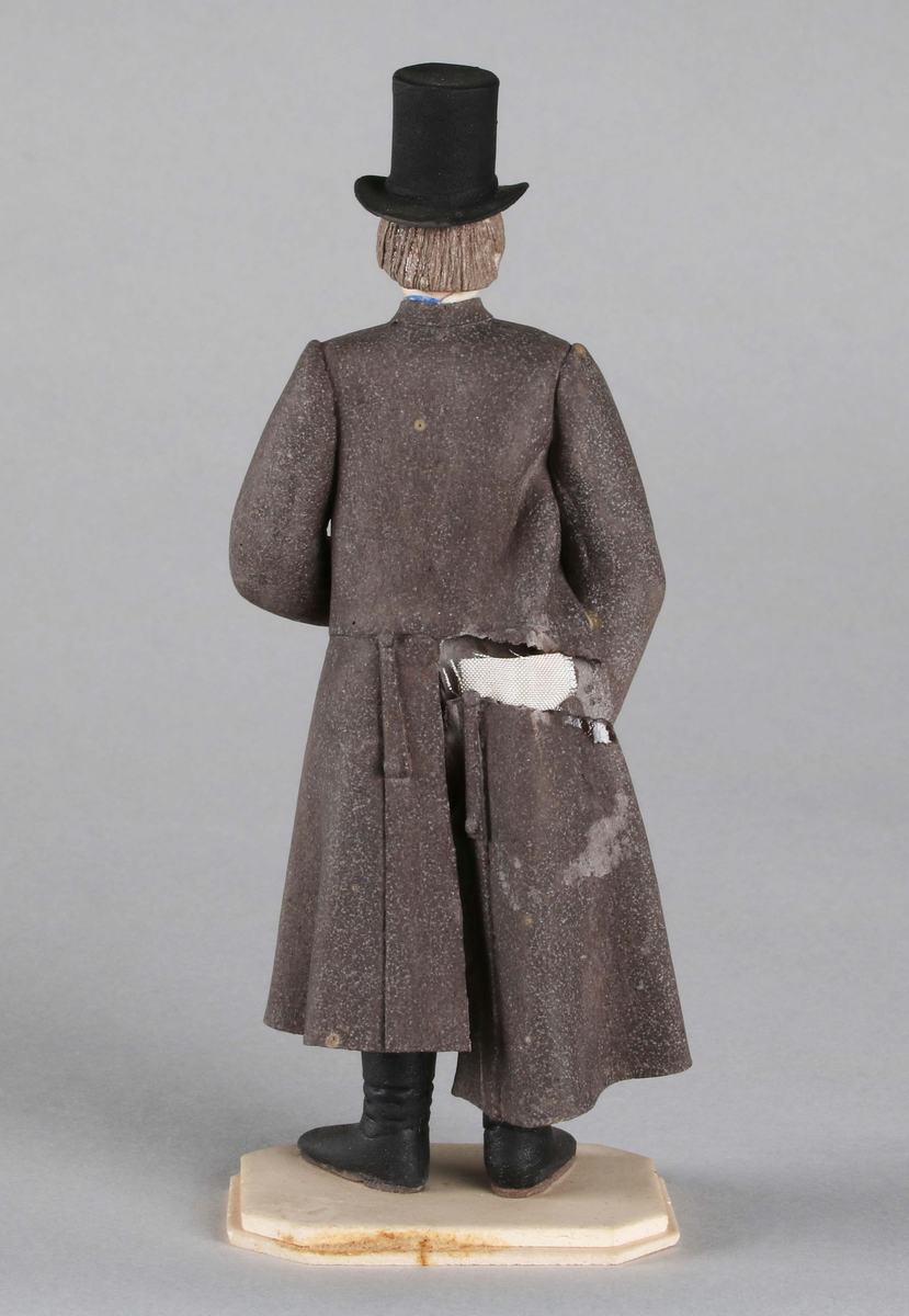Dragantarbete. Mansfigur.
Allmogedräkt. Stående mansfigur, iförd hög svart cylinderhatt. Trekvartslång, gråsvart rock med långa ärmar. Grön- och brunrandig väst och mörka knäbyxor. Svarta stövlar som räcka till knäna. Strumpor, mönstrade, blå och röda samt strumpeband. Blå knuten halsduk. Mannen bär en stor pipa i vänster hand. Står på åttkantig ljus platta.

Tillverkad av Hilda Boström (1822-1903), sockerbagare, Borås.

Litt: Se Årsbok 1963, sidan 45.

Funktion: Prydnadsfigur
