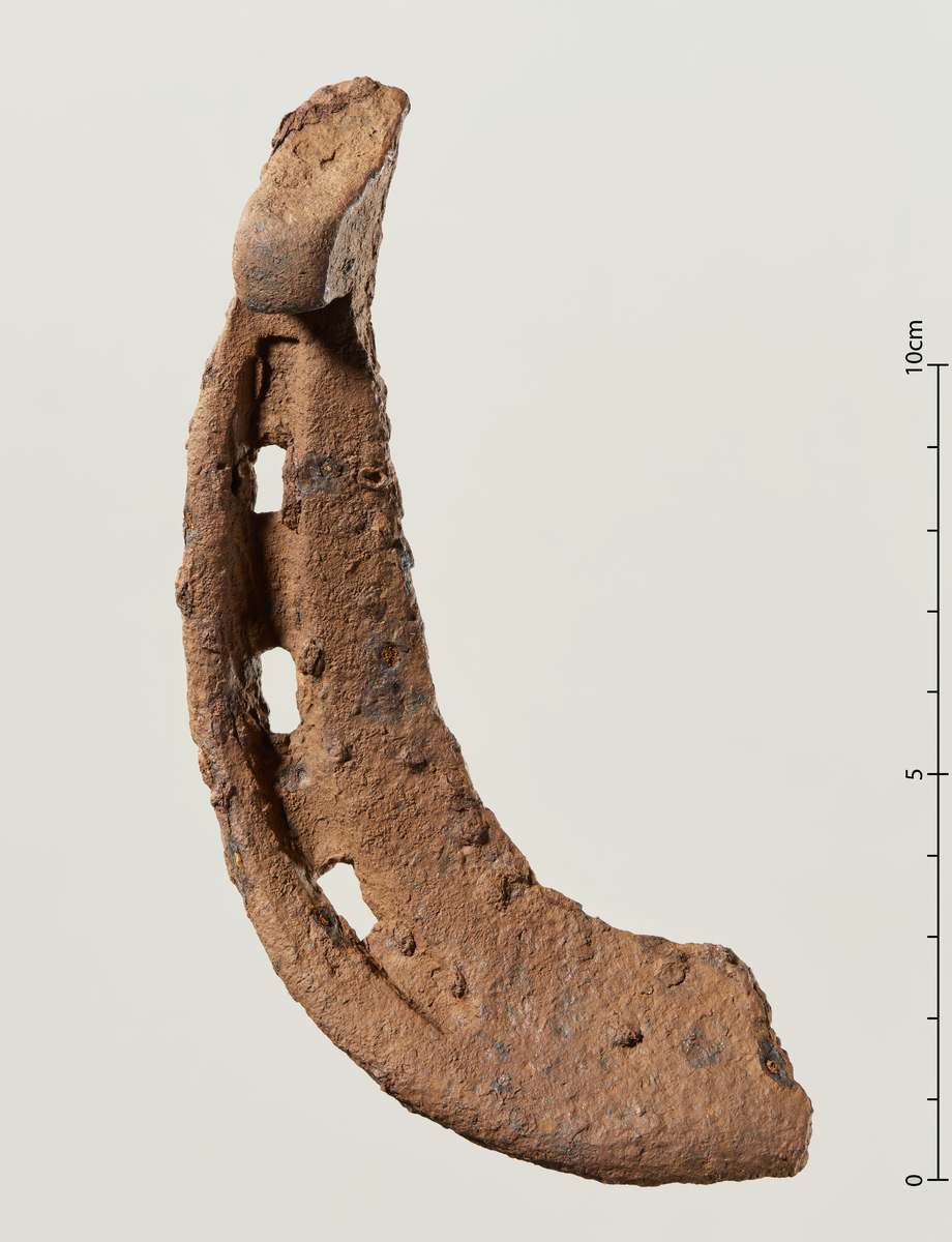 Fragment av hestesko med høye fremoverskrånende haker og reisning, som type C (Gansum 2002:32-33, Færden 1990:248). Halve skoen mangler, men fragmentet viser at den totalt har hatt seks rektangulære saumhull og vært ca 13,5 cm lang. Typologisk datert til mellom 1100- og 1400-tallet. 