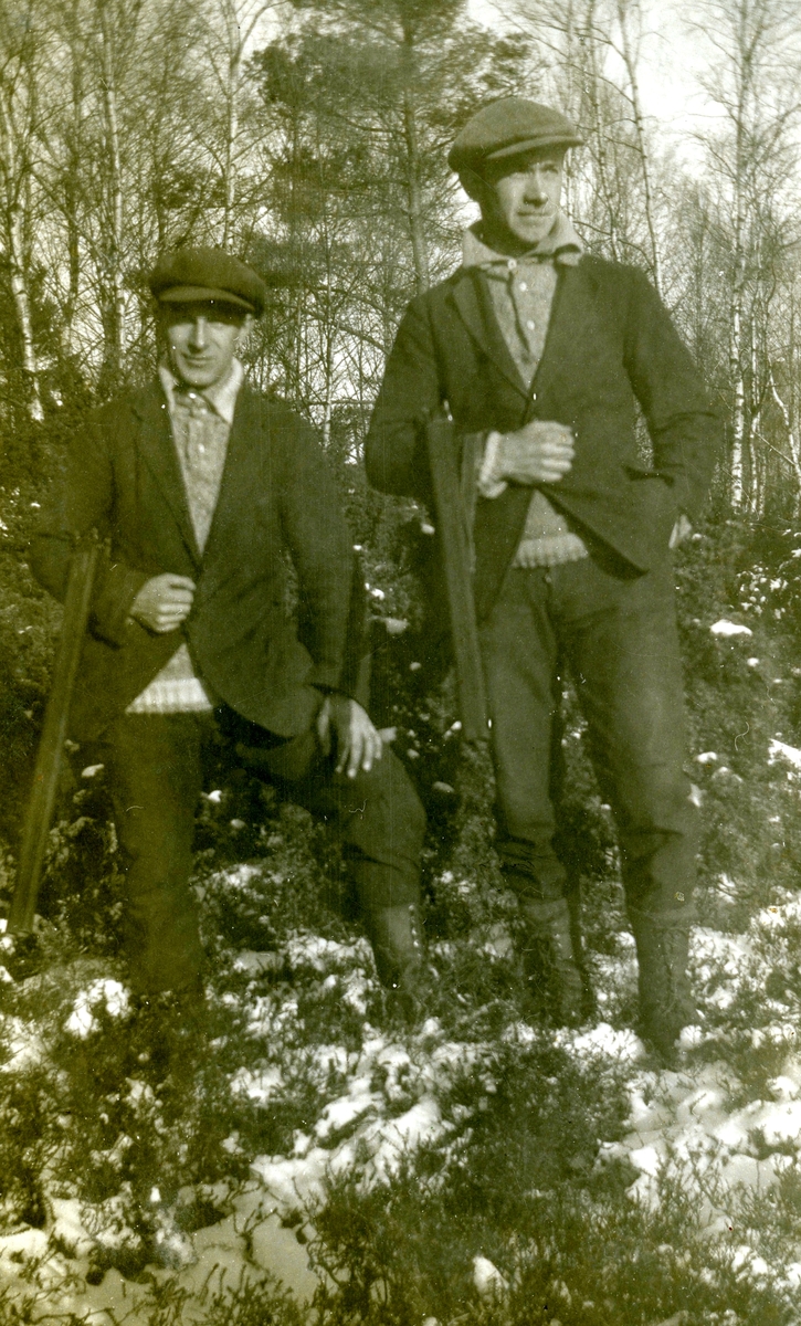 Två okända män, hållandes var sitt gevär, står på tomten vid Kållered Stom "Nygård" okänt årtal. På gräsmättan ligger lite snö.
