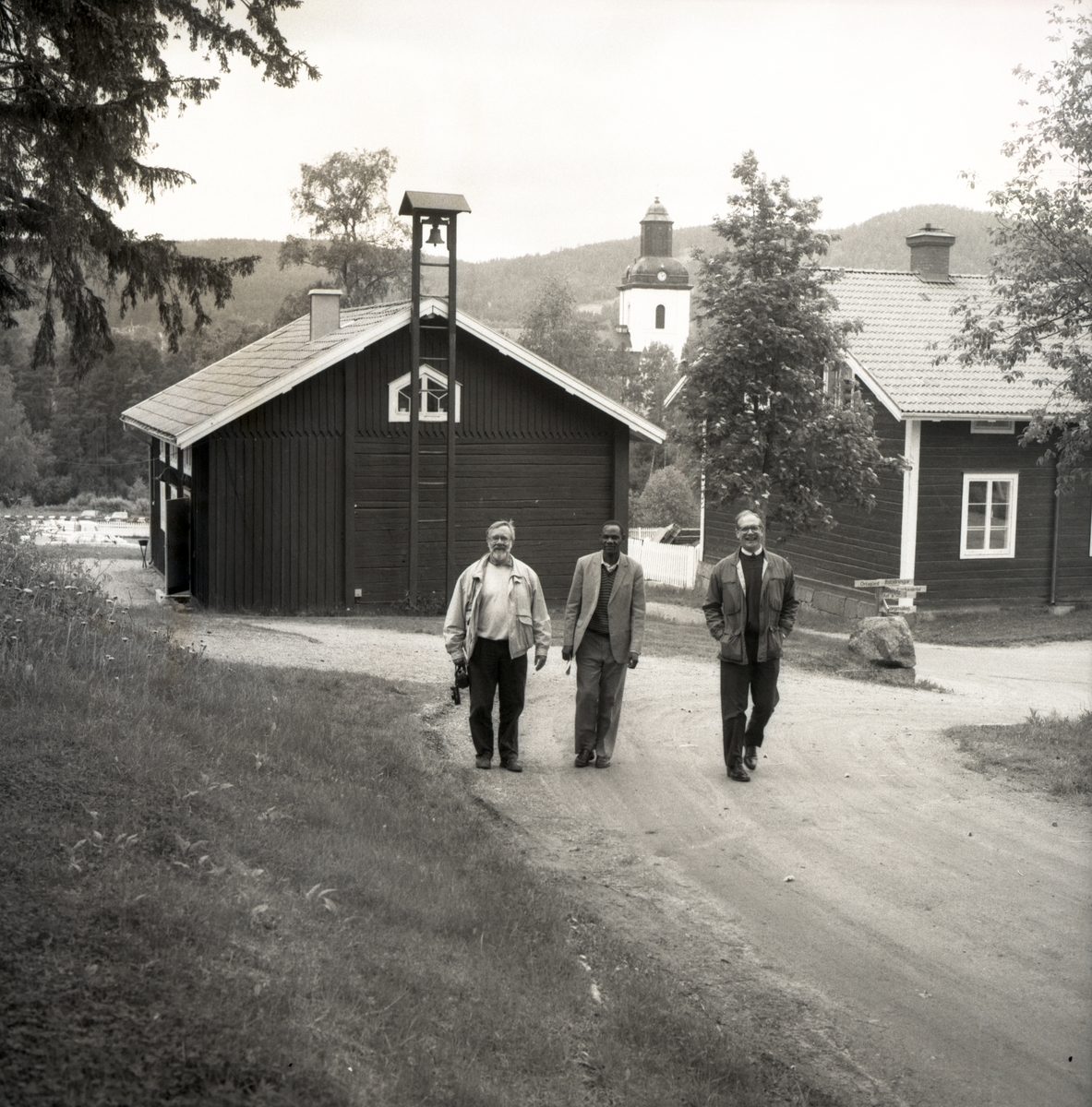 Tre män gående på en grusväg med en kyrka i bakgrunden. 1992.