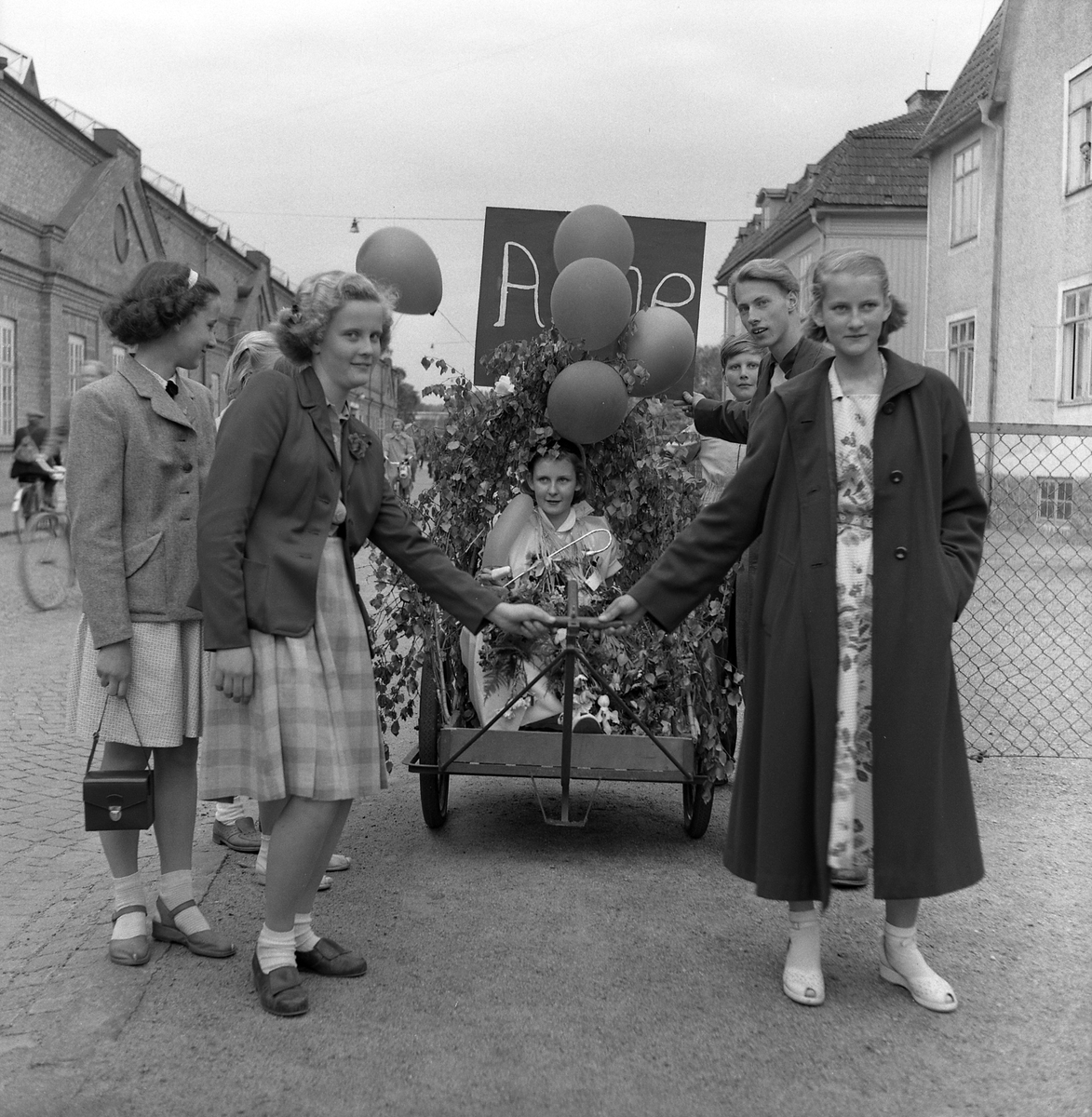 Ett antal personer kör en lövad vagn med en vän som tagit realen eller studenten. Från vänster: Anita Olofsson, okänd, Elisabeth Larsson (gift Danås), Anne (i vagnen), okänd, Bo Reteike, Rut (syster till Anne). Juni 1953