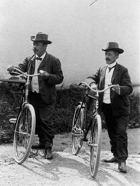 Två kostymklädda män med hatt står bredvid varsin cykel på en gårdsuppfart.
