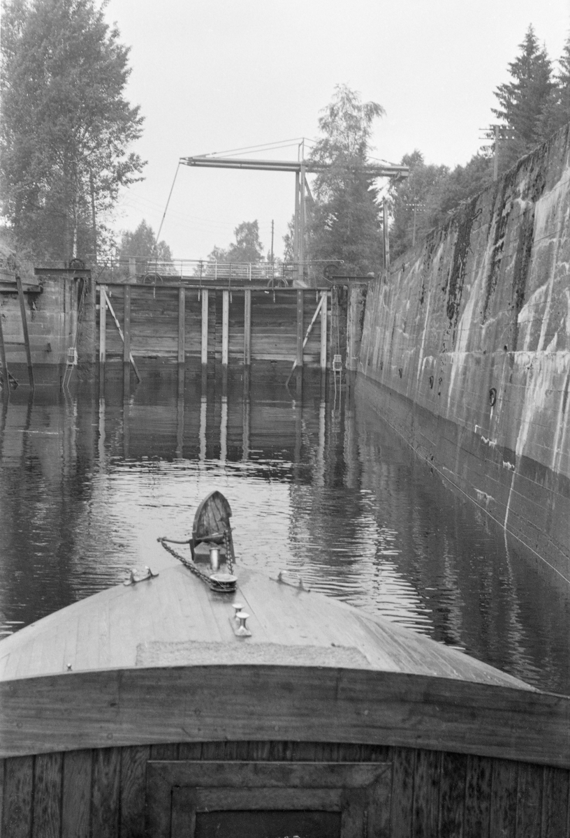 Fra Svanfoss sluse i Vorma, i Fenstad i Nes kommune i Akershus. Dette fotografiet ble tatt i 1954, fra en motorbåt som befant seg i slusekammeret, med kameraet vendt mot sluseporten på motstrøms side. Dam- og sluseanlegget ved Svanfoss ble bygd etter tegninger av daværende avdelingsingeniør i Kanalvesenet, seinere kanaldirektøt Ingvar Kristensensen (1865-1947). Anlegget skulle erstatte en eldre tømmerkistedam med sluse ved Sundfossen, cirka 13 kilometer høyere oppe i Vorma. I samråd med kanaldirektør Gunnar Sætren (1843-1928) foreslo Kristensen at slusa ved Svanfossen skulle bli 60 meter lang og 8 meter bred, med dybde på 3 meter ved lavvann. Ei slik dimensjonering skulle gjøre det mulig for dampfartøyer og flåter av buntet tømmer å passere dammen. 

Den 15. juli 1909 kunne avisa Akershus fortelle at en del nesbuer, på veg til et politisk møte på Eidsvoll, som de første hadde fått passere Svanfoss sluse: «Endel av Nesbuerne reiste med Udnes kvindeforenings motorbaat (Hagasundbaaten). Kl. 9,30 passertes sluserne ved Svanfos. Ingeniør Daae var saa venlig at aapne sluserne, dette var første gang noen gik gjennem der og første gang ruten mellem Haga og Eidsvold blev benyttet gjennem den nye, nu omtrent færdige kanal forbi Eidsvold.» Sommeren 1911 ble det igangsatt motobåtrute mellom Årnes og Eidsvoll med anløp også i Vormsund og ved Svanfoss sluse. Fra 1912 gikk motorbåten «Svan» på den samme strekningen, med atskillig flere stoppesteder