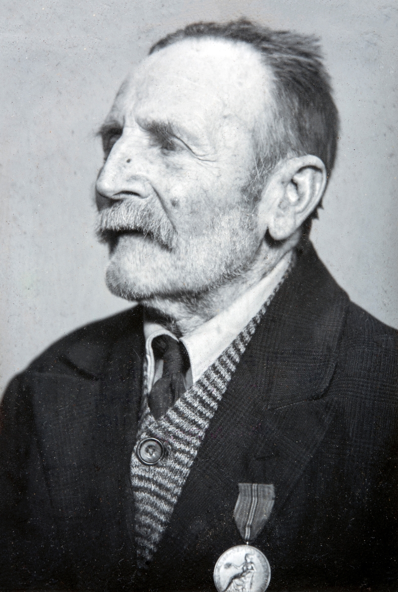 Portrett av Husmann Nils Olsen Hagen (29.01.1863-24.10.1955), Han var husmann på Hverven Gård i Ottestad, i rundt 60 år. På bilde har han medaljen for lang og tro tjeneste i fra Norges vel. Bildet er tatt i forbindelse med hans 90-års dag i 1953.