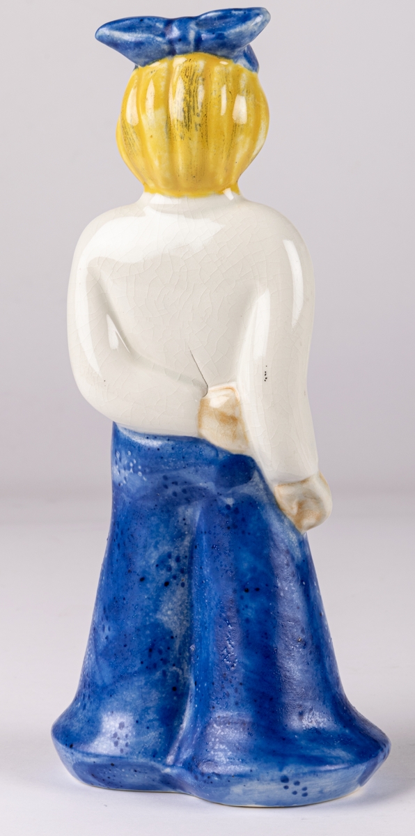 Figurin av flicka, Gretchen, blont hår, blå långbyxor, vit tröja och blå hatt, med ena handen på ryggen och den högra längs sidan. Handmålad. Formgiven 1937 av Märta Willborg.