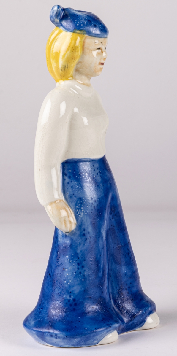 Figurin av flicka, Gretchen, blont hår, blå långbyxor, vit tröja och blå hatt, med ena handen på ryggen och den högra längs sidan. Handmålad. Formgiven 1937 av Märta Willborg.